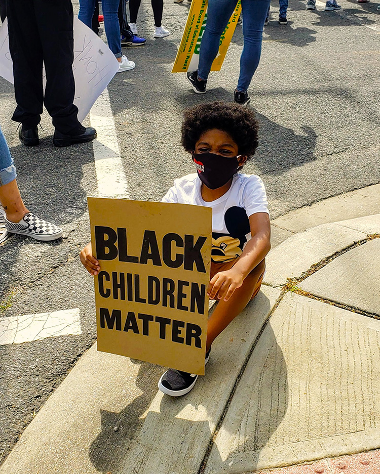 Cover photo for the Statement of Solidarity & Support. Shows a child sitting at a protest, holding a sign that reads "Black Children Matter"